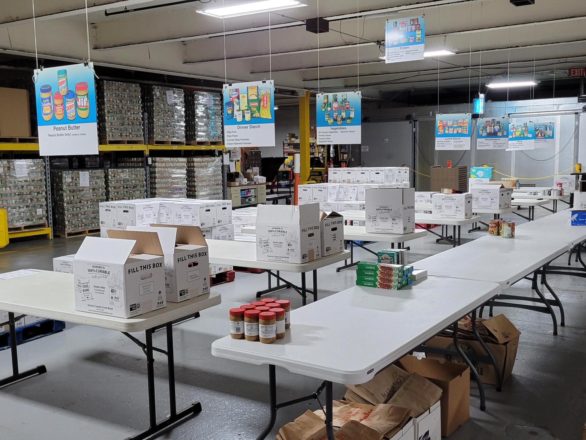 The Racine County Food Bank sorting area - several rows of white tables, each piled either with white Food Bank boxes or with stacks of household staples. Signs above the space label stations for different types of food, such as peanut butter and dinner starch. In the background, pallets full of cans are stacked to the ceiling of the warehouse.