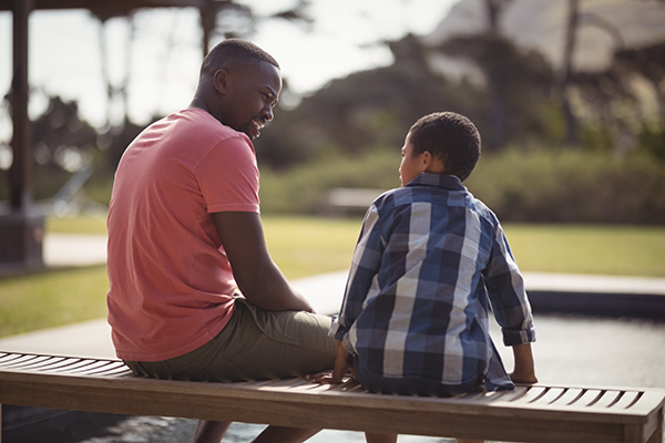 A Black adult and child sit beside each other on a bench, talking.