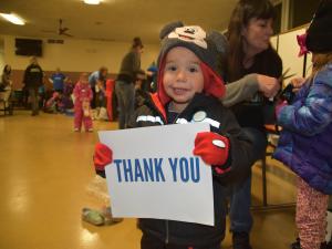 A toddler in just-donated winter clothes grins and holds a piece of paper that says "Thank you."