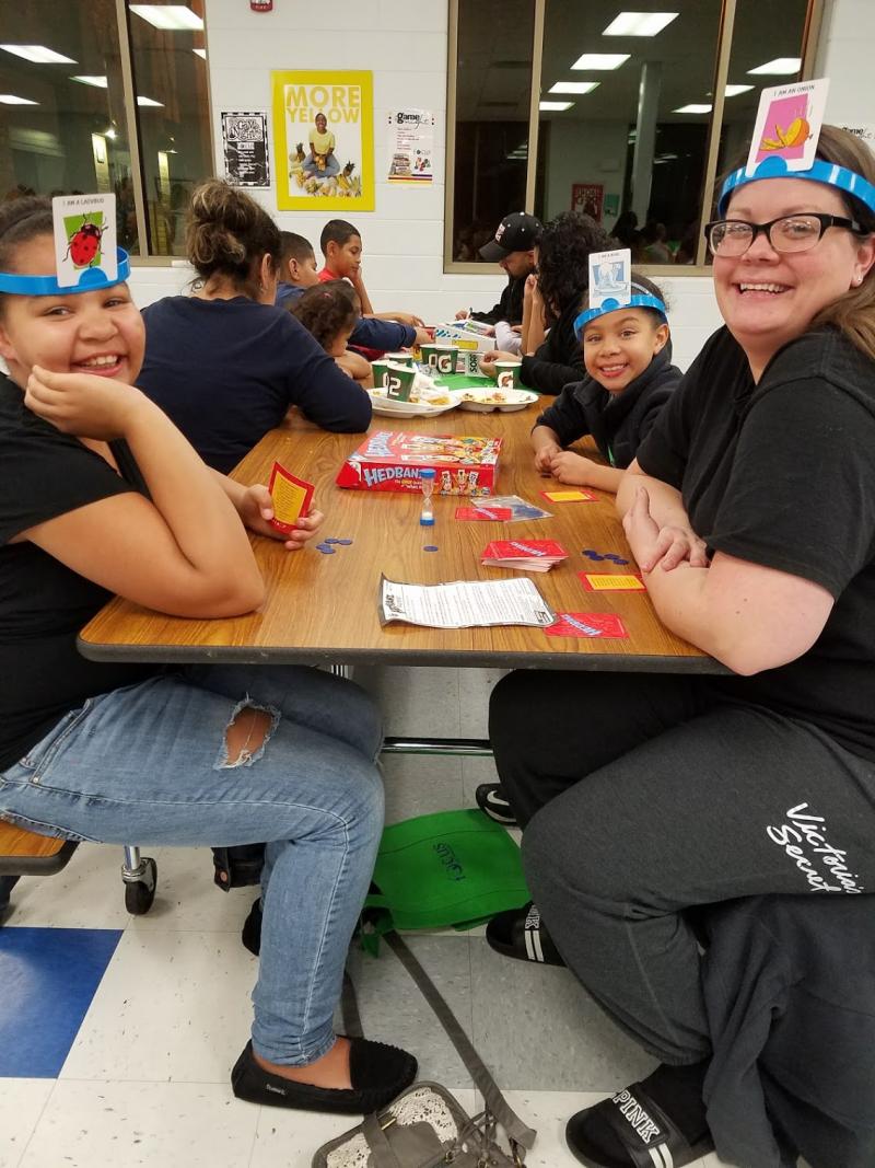 Kids and adults at a long table. The closest three people wear blue headbands with picture cards on the front, a game called Headbands. They grin at the camera.