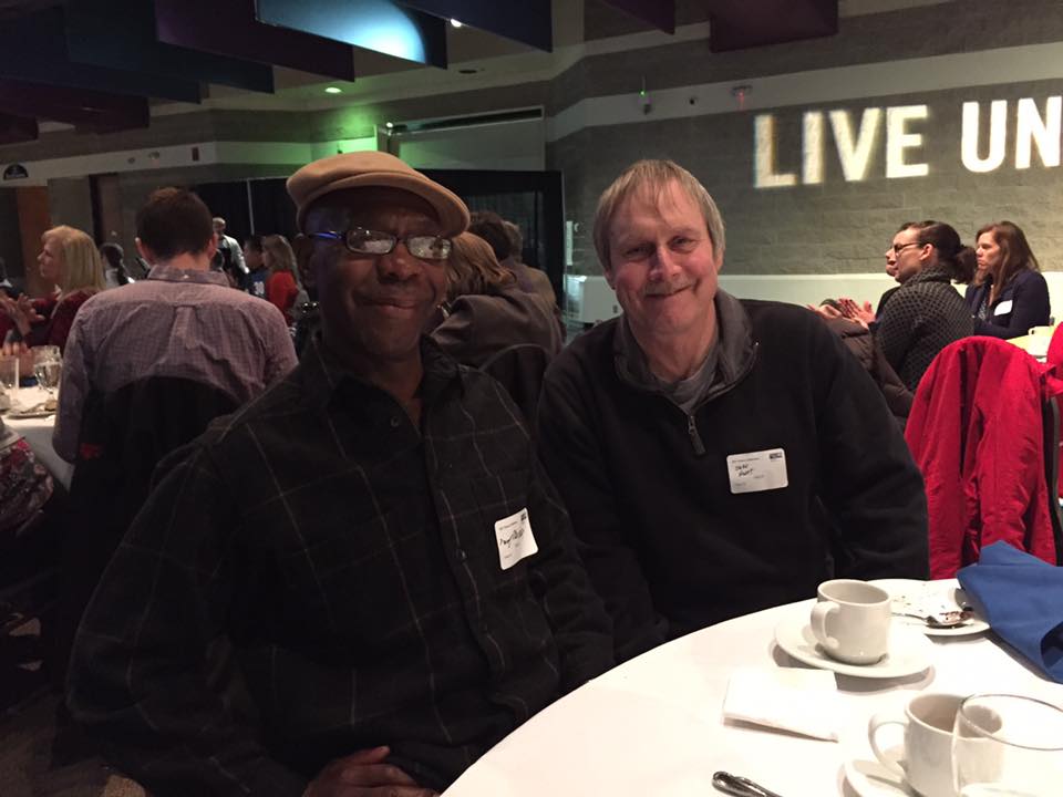Fred and Jeff, two former clients of HALO, share a table at United Way's Victory Celebration. They lean together and grin at a photographer. "Live United" is projected in white light on the wall behind them.