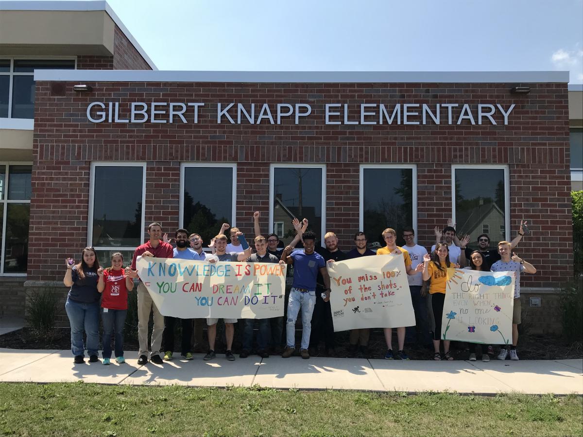 A group of adults stand under the Gilbert Knapp Elementary sign on the side of the school. They hold inspirational banners and raise their hands in the air.