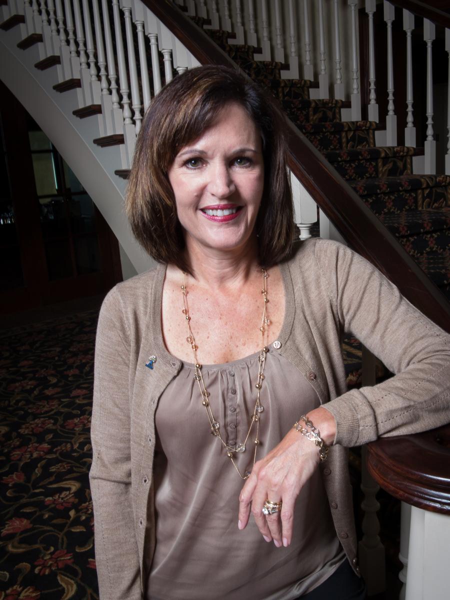 IMAGE DESCRIPTION. Liz Powell, executive director of the Racine Community Foundation, leans against a staircase and smiles at the camera. Her long, brown bob just barely reaches her shoulders. She wears a tan blouse and tan cardigan with a long, dangling necklace. Her pale, raised wrist has a thin, silver bracelet. END IMAGE DESCRIPTION.