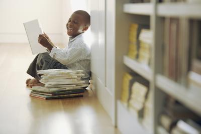An elated child holds a book, sitting next to a pile of books in front of a shelf of books.