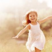 A photo of a thin, white child with long hair and a white dress. They run through a misty field with a big grin and their arms spread wide.