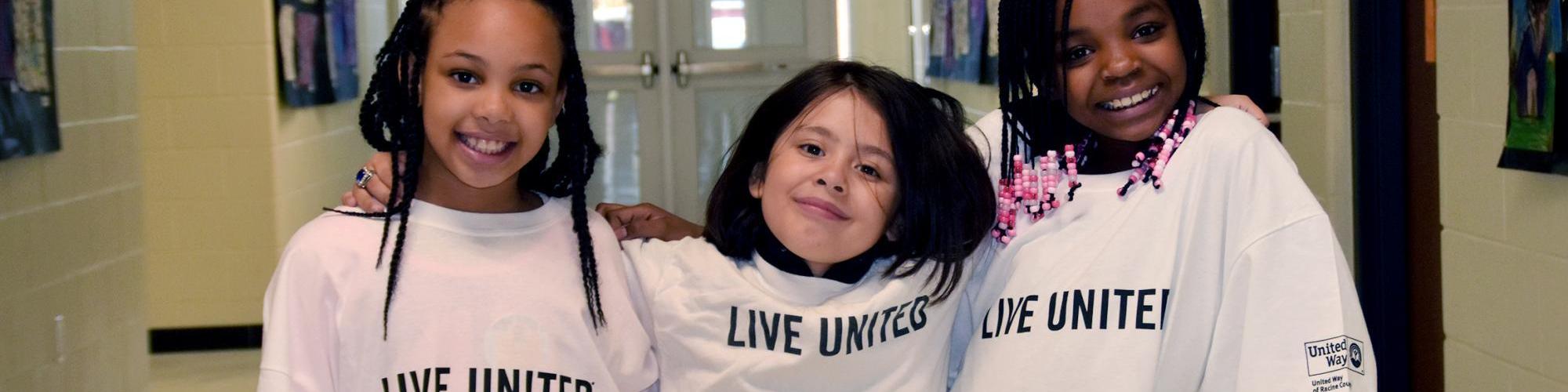 Three kids in the hall of Knapp Elementary wearing oversized Live United shirts wrap their arms around each other's shoulders and strike cool poses for the camera.