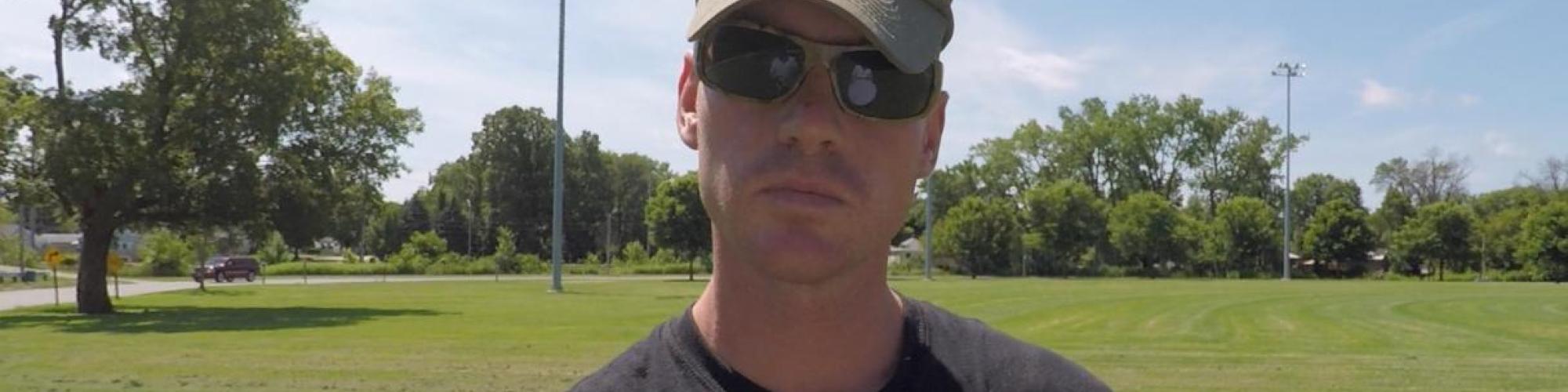 A photo of David staring at the camera with a neutral expression, head slightly tilted and eyes concealed by sunglasses and the brim of a cap that says "Iraq Afghanistan Veteran."