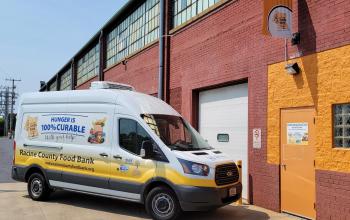 The Racine County Food Bank's van is parked at an angle in front of its front entrance - an orange-painted door and section of wall on a red brick warehouse building.