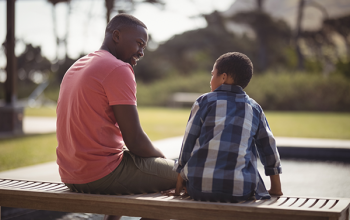 A Black adult and child sit beside each other on a bench, talking.
