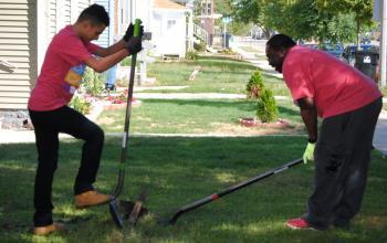 Photo of two Day of Caring volunteers landscaping at the George Bray Neighborhood YMCA. Their shovels pierce the earth at the same place, one volunteer bending down and the other with their foot perched on the shovel.