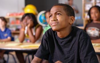A kid squints up from their desk at school. Blurred out behind them, other students sit in various poses of focus.