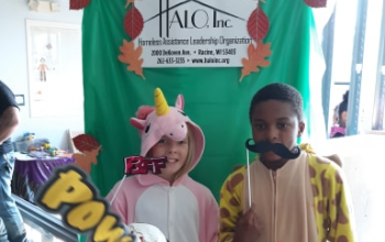 Three kids living at HALO wear Halloween costumes and pose. One wears a unicorn onesie and holds a "BFF" sign on a stick; one wears a giraffe onesie and holds a mustache on a stick to their mouth; and in front of them both, a clown wearing a half-mask of a bloody skull holds up "pow" and "booom" signs. Behind the kids is a green backdrop with the HALO logo.