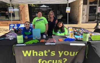 Four smiling kids and adults stand behind a table with a tablecloth that says "What's your focus?" The table is covered in wristbands, lollipops, kindness rocks, donation jars and other materials.