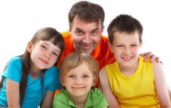 Three kids sit in front of their parent, who has a hand on the shoulder of one of the kids. They're all wearing different colored shirts and smiling.