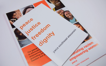 A pile of YWCA collateral on a white background. The top piece says "peace, justice, freedom, dignity. ywca southeast wisconsin." The bottom piece, which is partially obscured by the top piece, has various pictures of Black people and YWCA's name and mission statement.