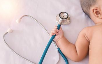 Photo centers on a stethoscope, which a baby partially off camera grips in their right hand. Both baby and stethoscope are nestled on a fluffy, white layer of fabric.