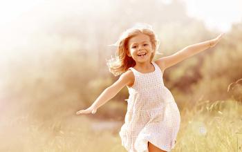 A photo of a thin, white child with long hair and a white dress. They run through a misty field with a big smile and their arms held out wide.